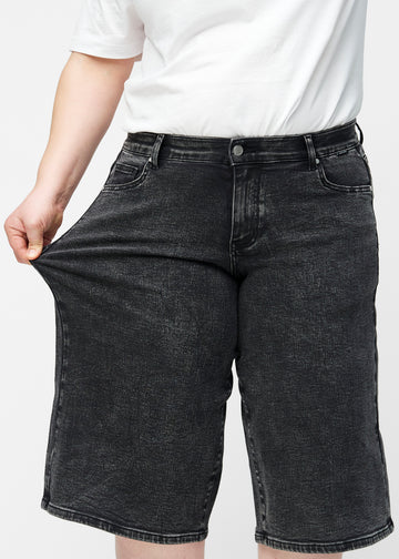 Plus-size model strækker de løse, mørkegrå denim capris i farven Shadows ved hoften for at vise materialets stretch.