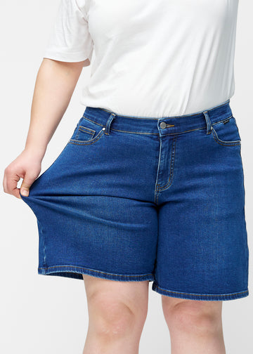 Plus-size model strækker de løse, mørkeblå denim shorts i farven Oceans ved hoften for at vise materialets stretch.