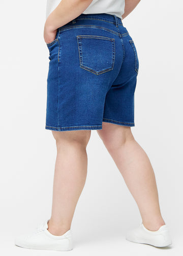 Løse, mørkeblå denim shorts i farven Oceans set fra siden på plus-size model, hvor den brede pasform og længden til lige over knæet er synlig.