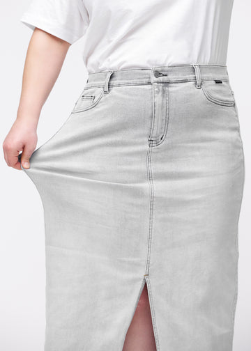 Plus-size model strækker den lange, lysegrå denimnederdel i farven Ashes ved hoften for at vise materialets stretch.