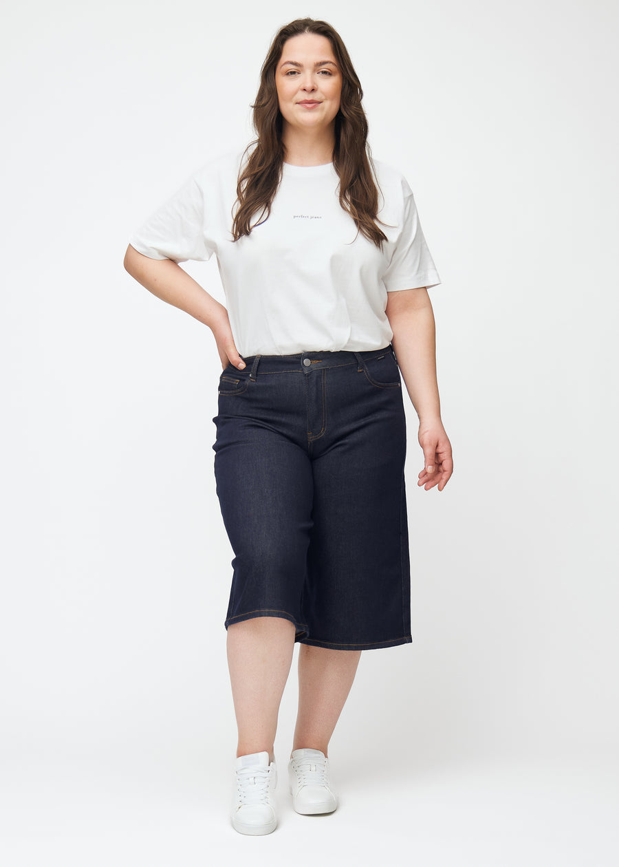 Full image of a plus-size model wearing deep dark blue denim capris for women in the colour Midnights with a loose fit.