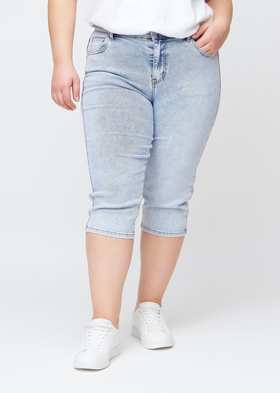 Slim capris for women in light blue denim, color Waves. Front view on a plus-size model, showing the fitted cut ending just below the knee.
