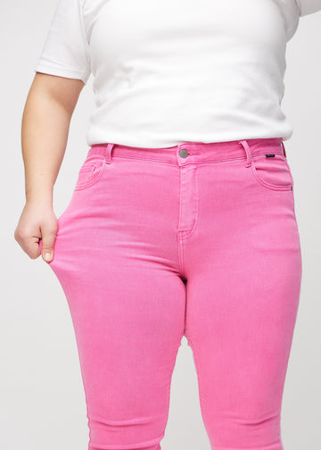 Skinny jeans for women in light pink, color Flamingos. A plus-size model stretches the jeans at the thigh to show the fabric&