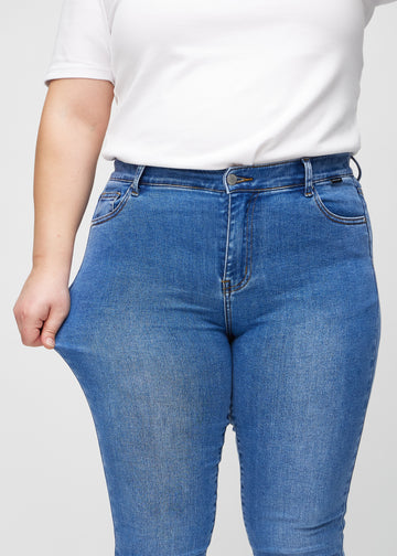 Skinny jeans for women in blue, color Rivers. A plus-size model stretches the jeans at the thigh to show the fabric&