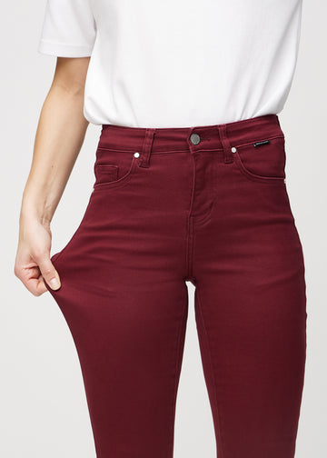 Skinny jeans for women in dark red, color Santas. A model stretches the jeans at the thigh to show the fabric&