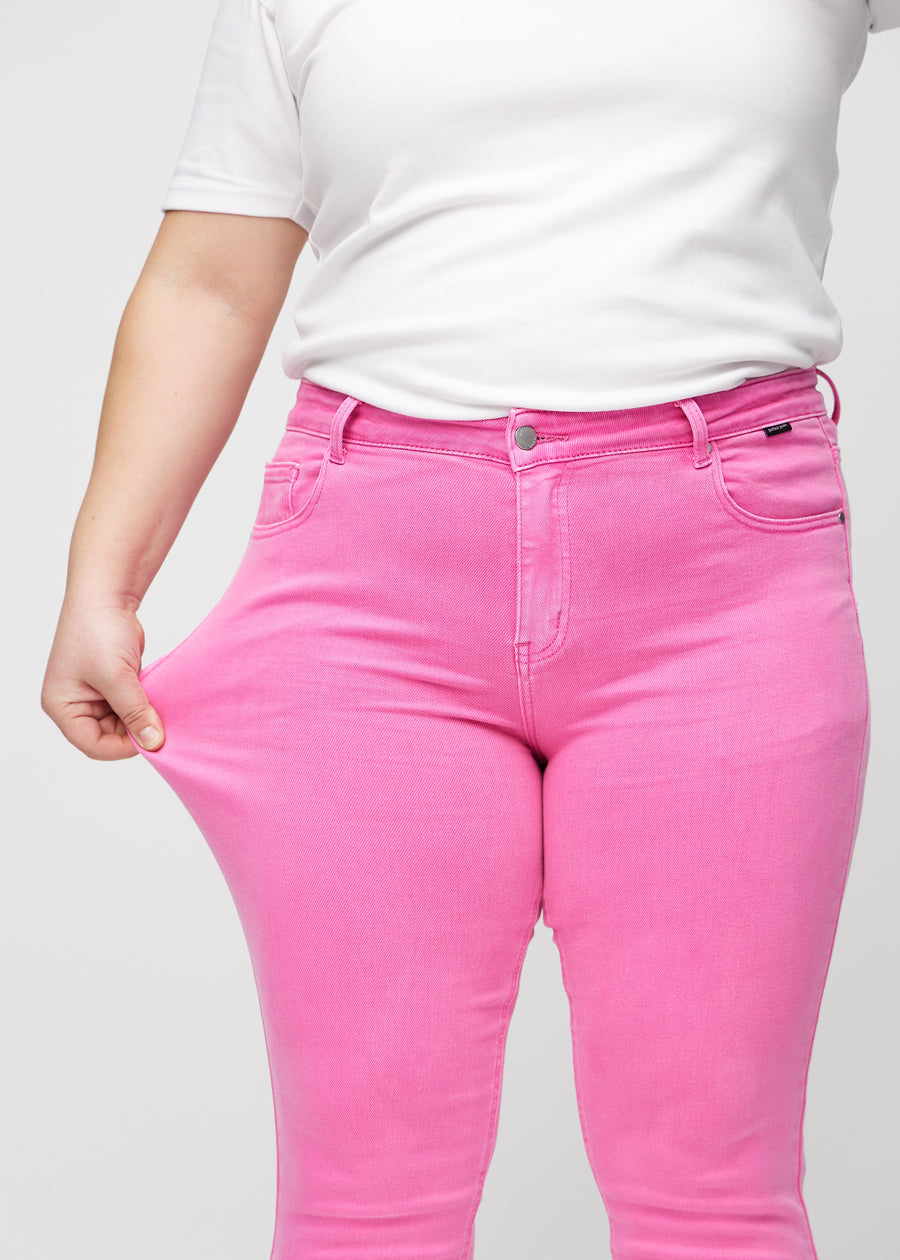 Slim jeans for women in light pink, color Flamingos. A plus-size model stretches the jeans at the thigh to show the fabric&