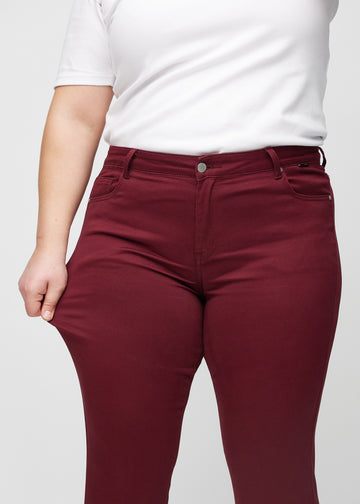 Slim jeans for women in dark red, color Santas. A plus-size model stretches the jeans at the thigh to show the fabric&