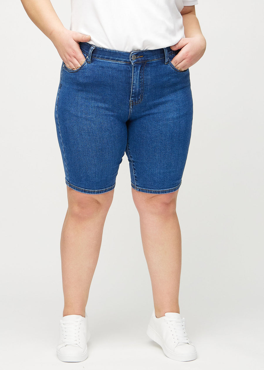 Dark denim skinny middle shorts, model name Oceans, which fit closely around the thigh and end just above the knee on a plus-size model, seen from the front.